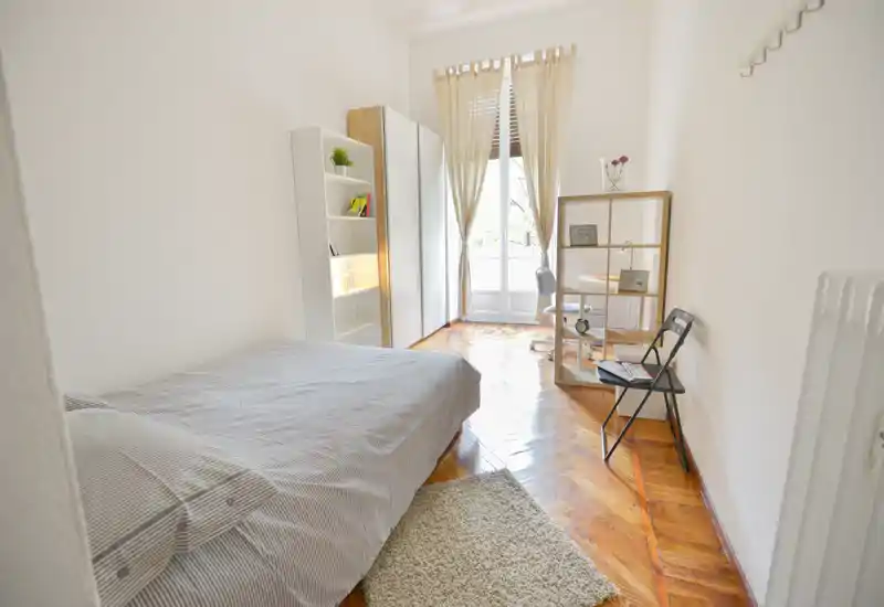 Camera da letto luminosa e ariosa con comodo letto matrimoniale, pavimento in parquet e piccolo angolo studio vicino alla finestra. Lenzuola neutre e scaffalatura semplice creano un ambiente ordinato e rilassante.