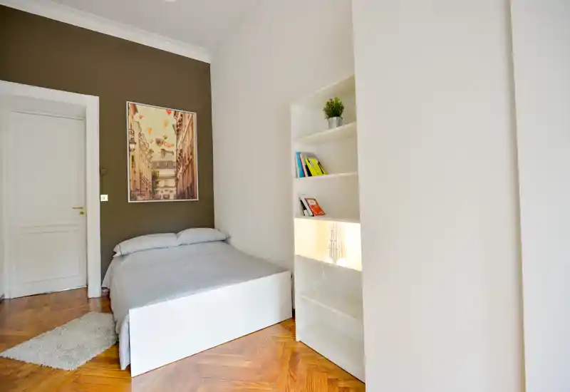 Camera da letto luminosa e minimal con letto matrimoniale, biancheria grigia e scaffalatura incorporata; pavimento in parquet e tonalità neutre creano un ambiente accogliente.