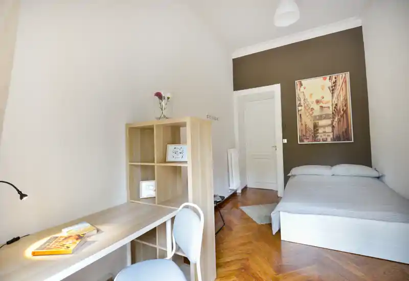 Camera da letto luminosa e ordinata con letto singolo, scrivania e scaffalatura: ideale per uno studente o un occupante singolo. Il pavimento in legno caldo e il quadro creano un'atmosfera tranquilla e accogliente.