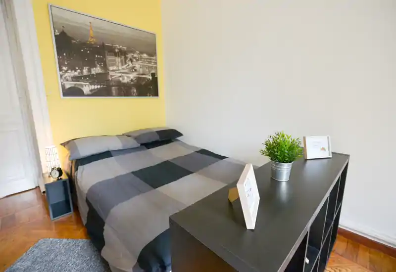 Chambre lumineuse et soignée avec un lit double en nuances de gris, un cadre décoratif et une étagère basse faisant office de séparateur — idéale pour une annonce.