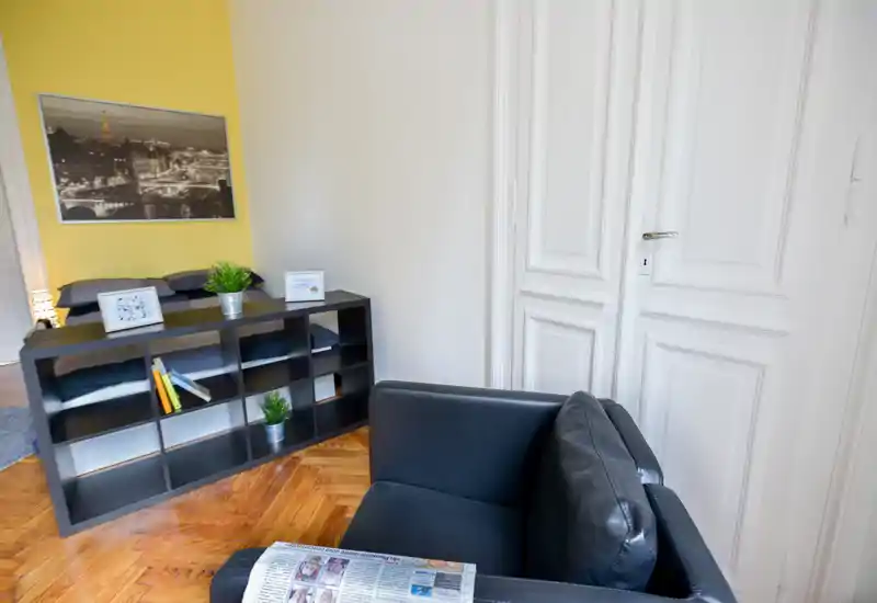 Coin chambre lumineux et moderne avec un lit double visible derrière une étagère basse, des plantes décoratives et un fauteuil noir au premier plan.