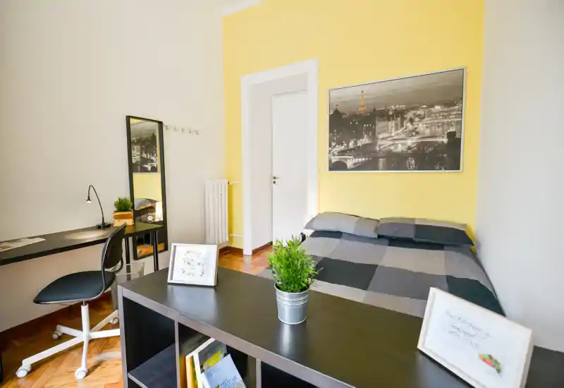 Chambre lumineuse et bien mise en scène avec lit double contre un mur jaune, tableau urbain, bureau et étagères — idéale pour les locations de courte durée ou un séjour confortable.