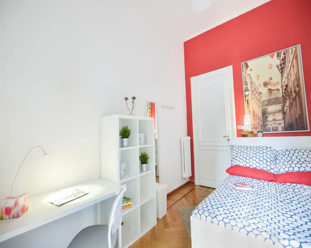 Camera da letto moderna e luminosa con trapunta a motivi, parete rossa e un piccolo angolo lavoro ordinato. Presenti letto matrimoniale, lampada, quadro e scaffale, perfetta per soggiorni confortevoli e smart working.