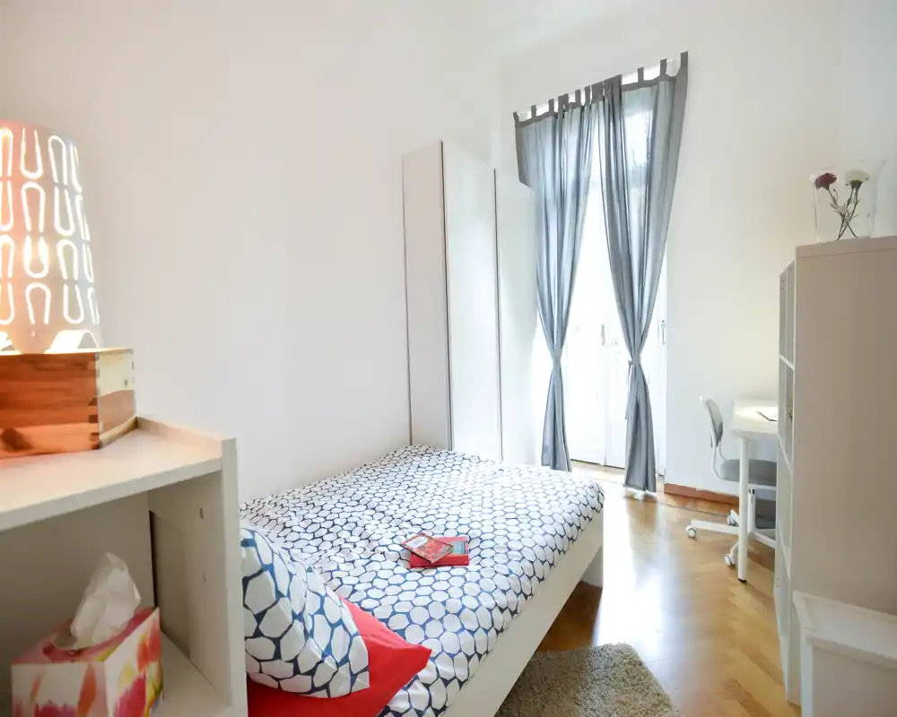Camera da letto luminosa e ben arredata con letto matrimoniale, biancheria a motivo, tappeto morbido e scrivania vicino alla finestra — ideale per una persona o una coppia. La luce naturale attraversa le tende alte creando un’atmosfera accogliente.
