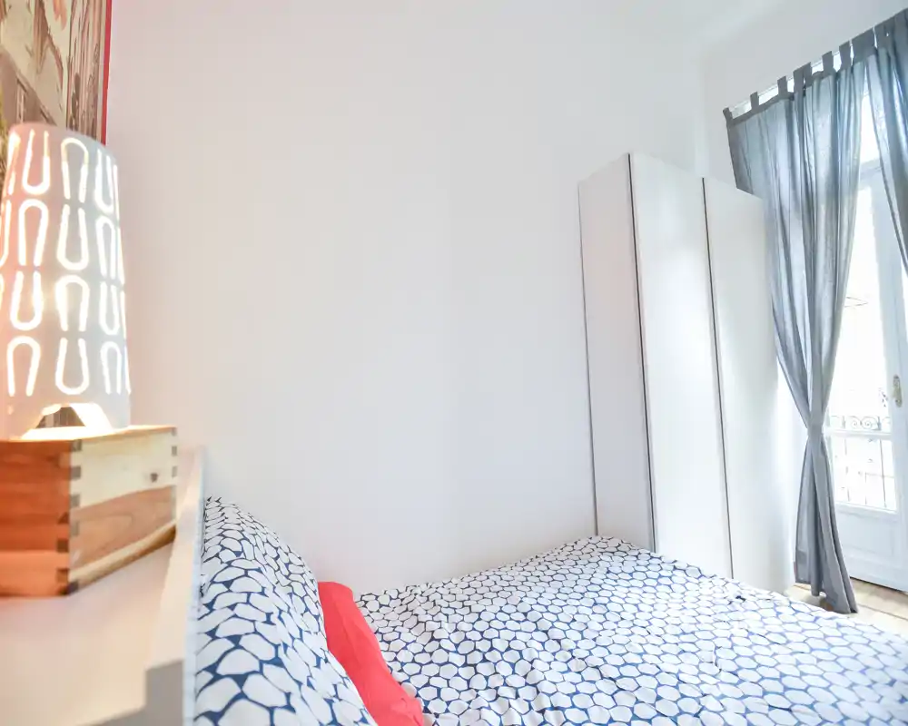Camera da letto luminosa e moderna con biancheria a fantasia, lampada sul comodino e un armadio bianco accanto alla finestra con tende leggere — uno spazio accogliente e ben illuminato.