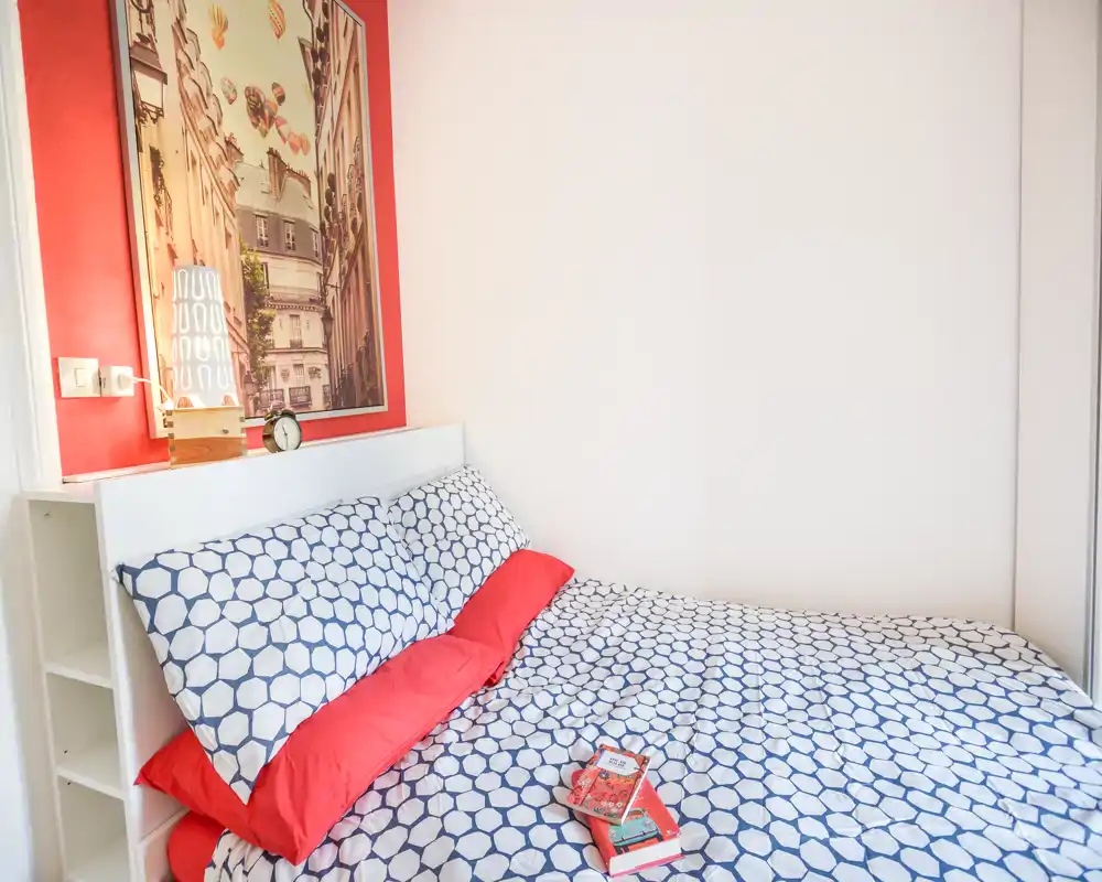 Camera da letto luminosa e accogliente con copripiumino blu e bianco a motivo, cuscini rossi e testiera semplice; scaffalatura laterale e quadro completano l’ambiente.
