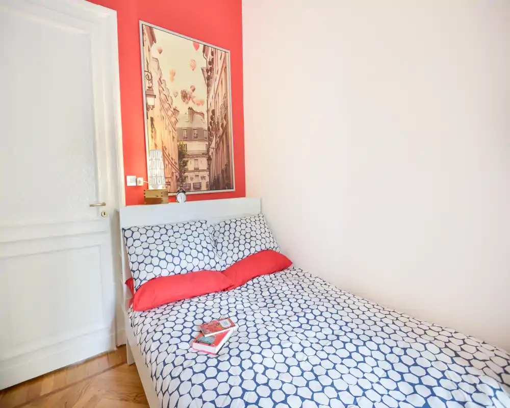 Camera da letto luminosa e compatta con letto matrimoniale, copertura a motivo e cuscini rossi; un quadro e una mensola completano l'ambiente accogliente.