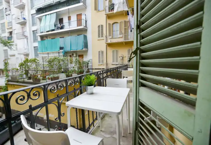 Balcone stretto e soleggiato con piccolo tavolo bianco, due sedie e una pianta in vaso che si affaccia sugli edifici vicini — ideale per il caffè mattutino.