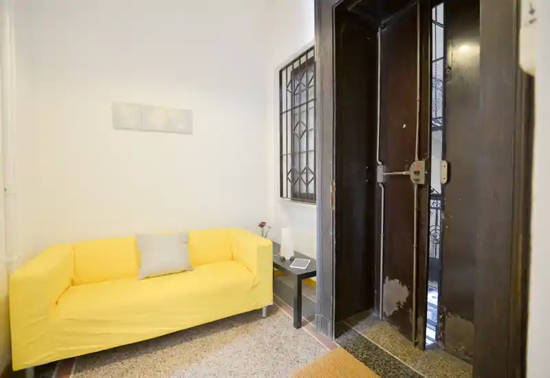 Piccolo ingresso con divano giallo brillante, tavolino laterale e una porta interna robusta; spazio pulito e ben illuminato adatto come area d'accoglienza.