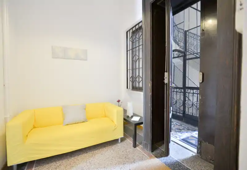 Piccolo soggiorno con divano giallo brillante, tavolino e porta aperta che dà su una scala in ferro decorata. Spazio pulito e ben illuminato ideale per un soggiorno compatto.
