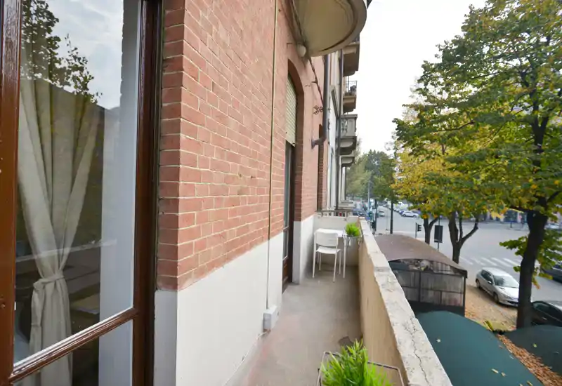 Balcone urbano stretto con passaggio, piccolo tavolo e sedie e vista sulla strada circondata da alberi maturi — perfetto per un caffè mattutino.