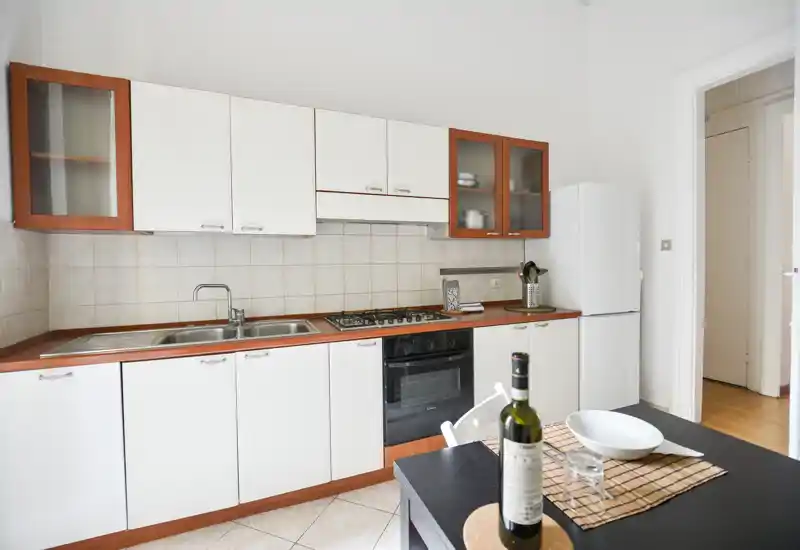 Cucina luminosa e ben attrezzata con mobili bianchi, lavello integrato e piano cottura a gas, più un piccolo tavolo apparecchiato con bottiglia e ciotola — perfetta per preparare pasti in sicurezza.