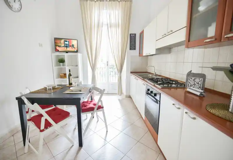 Cucina compatta e luminosa con piano cottura e forno integrati, pensili bianchi e un piccolo tavolo da pranzo con sedie con cuscini rossi — un ambiente accogliente ideale per un appartamento cittadino.