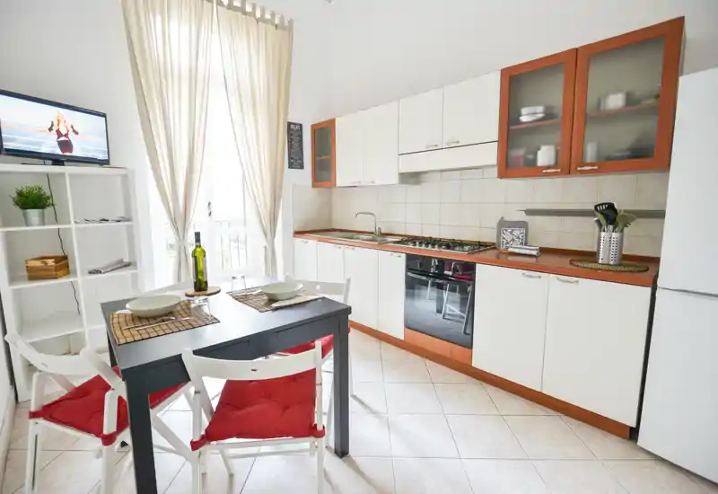 Cucina compatta e luminosa con forno integrato, piano cottura a gas e un piccolo tavolo da pranzo per due; tende chiare e pensili con dettagli in legno creano un’atmosfera accogliente.