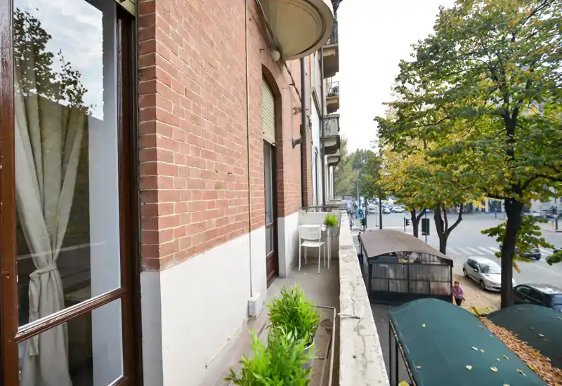 Balcone stretto e soleggiato con piante in vaso, piccolo tavolo e sedie che si affacciano su una strada alberata — perfetto per il caffè mattutino.