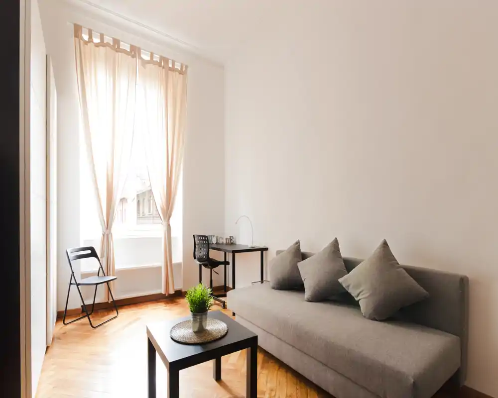 Bright, minimalist living room with a grey sofa, throw cushions, a small coffee table with a plant, and a desk by a large window letting in soft natural light.