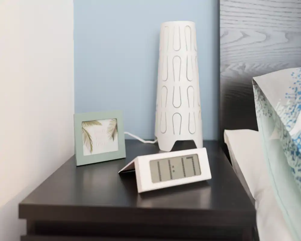 Close-up of a bedroom bedside table with a modern lamp, small digital clock and decorative photo frame next to the edge of the bed.