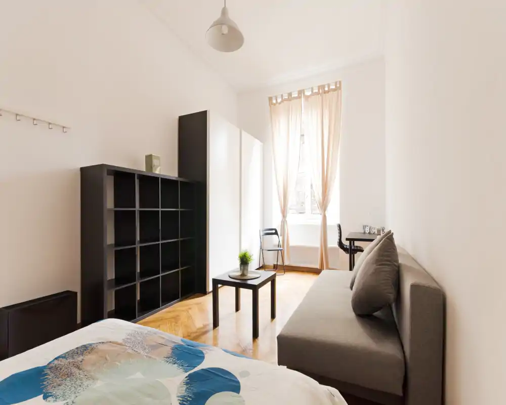 Bright, minimalist bedroom with a comfortable bed in the foreground, a grey sofa, shelving unit and a window with soft curtains that floods the room with natural light — ideal for modern rental listings.