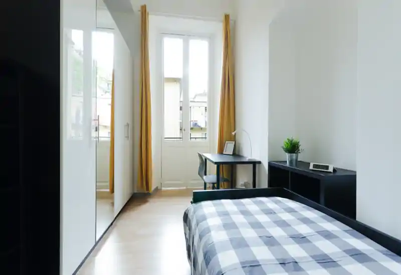 Chambre simple lumineuse et moderne avec lit bien fait, bureau près de la fenêtre et rangement minimaliste — parfaite pour un étudiant ou une personne seule.