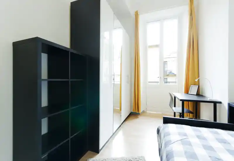 Chambre moderne et lumineuse avec lit bien fait, garde-robe à portes miroir, étagères et bureau près de grandes fenêtres avec rideaux jaunes apportant une lumière naturelle chaleureuse.