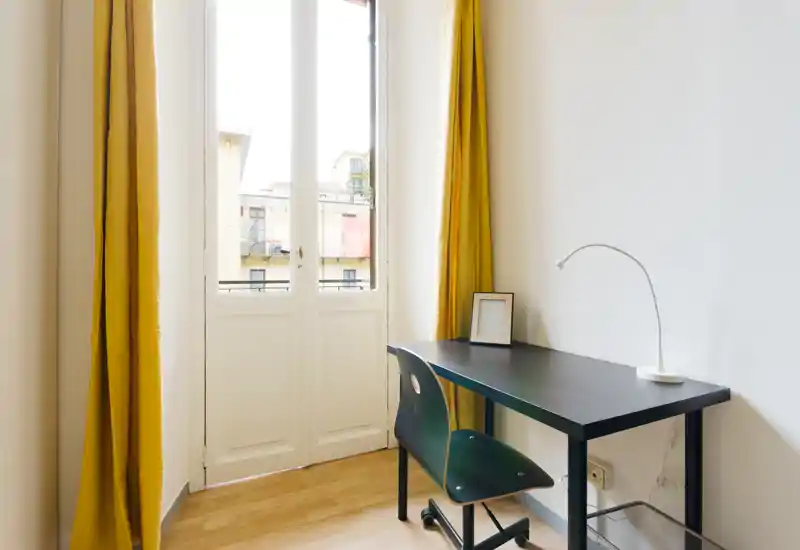 Coin bureau lumineux avec un bureau noir simple, une chaise et une lampe près d'une grande fenêtre avec rideaux jaunes — idéal pour travailler ou étudier.