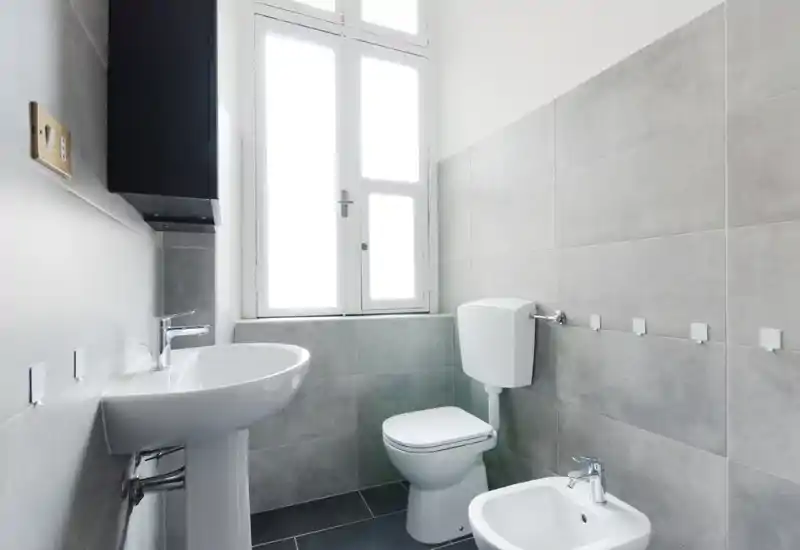 Bagno moderno e luminoso con lavabo a colonna, WC, bidet e ampia finestra che illumina naturalmente; piastrelle grigie neutre e design minimalista pulito.