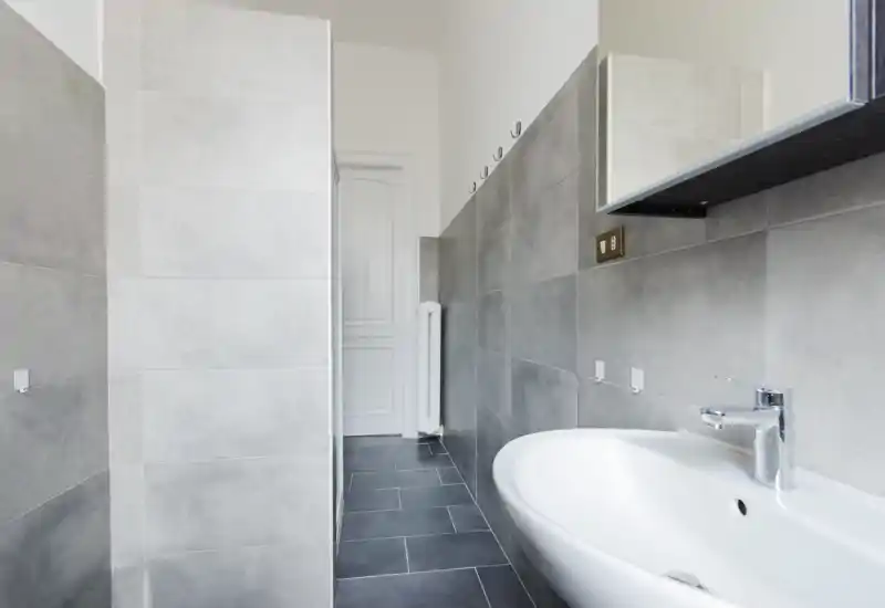 Bagno moderno con piastrelle grigie, lavabo bianco e rubinetto cromato, e area doccia a vista in un layout pulito e contemporaneo.