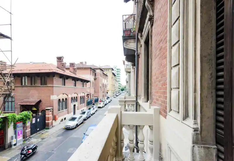 Piccolo balcone in pietra che si affaccia su una tranquilla strada cittadina con auto parcheggiate e palazzi in mattoni storici; luce diurna e toni neutri adatti per annunci.