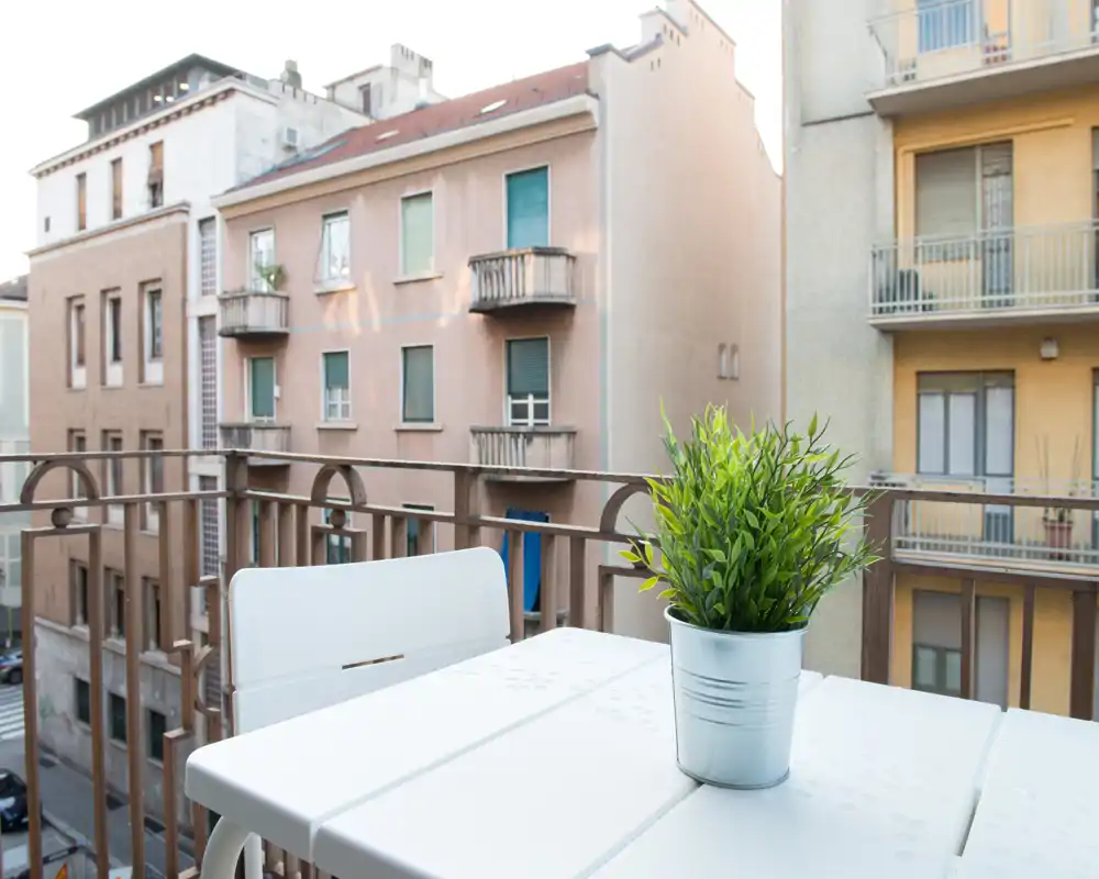 Petit balcon ensoleillé avec table blanche, chaise et une plante en pot donnant sur les immeubles voisins — idéal pour un café ou une pause.