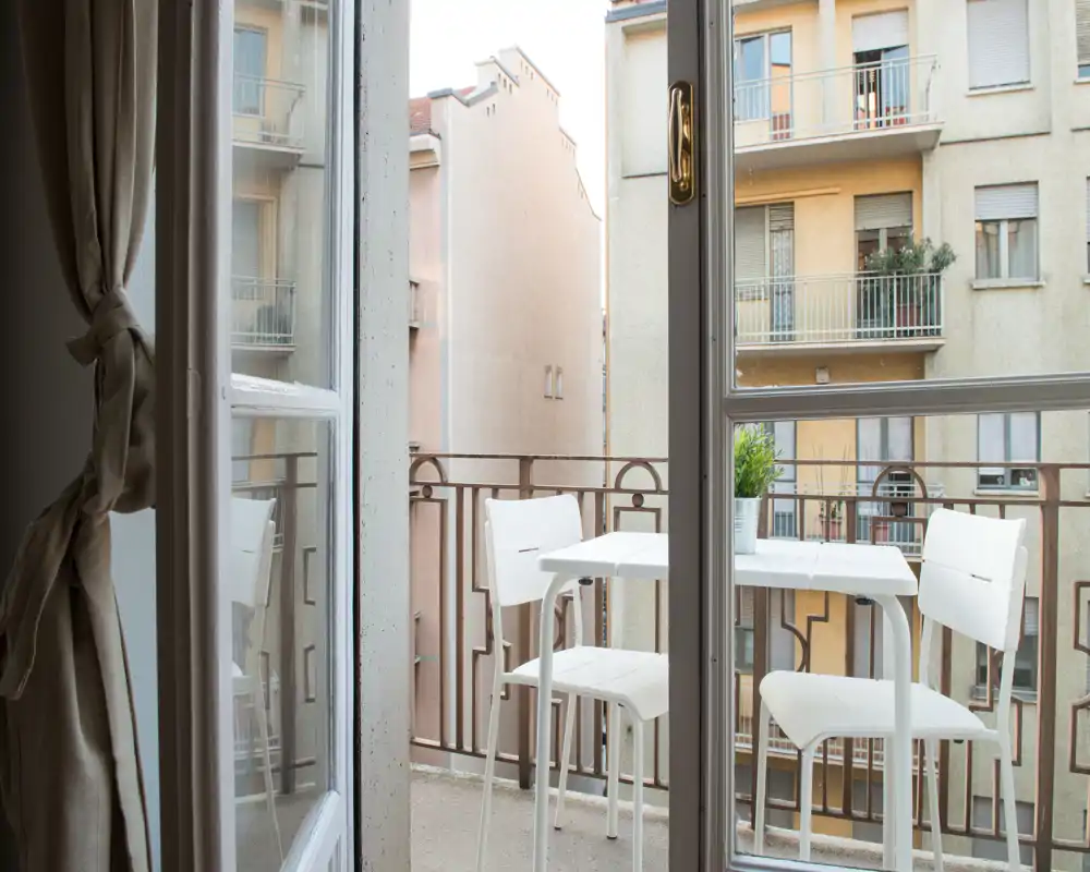 Petit balcon en ville avec une table blanche compacte et deux chaises, lumière naturelle vive et vue sur les façades des immeubles voisins.