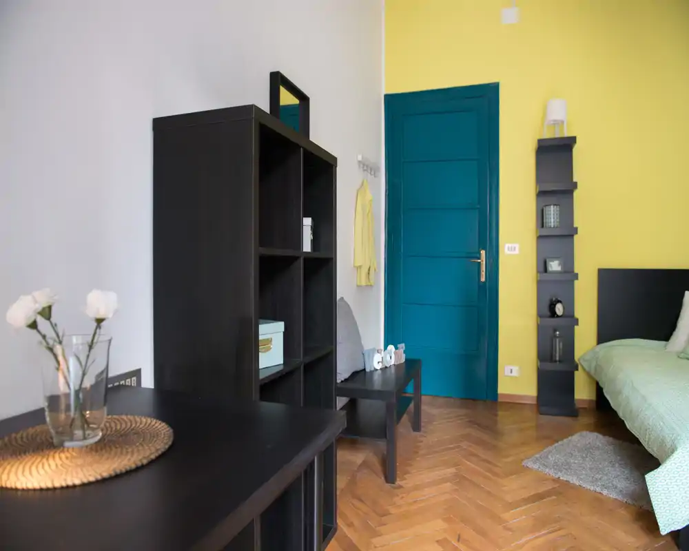 Coin chambre lumineux et moderne avec une porte bleu sarcelle et un mur jaune contrastant. Le lit est visible à droite avec une literie verte, complété par des étagères foncées et un parquet en bois pour une ambiance chaleureuse et contemporaine.