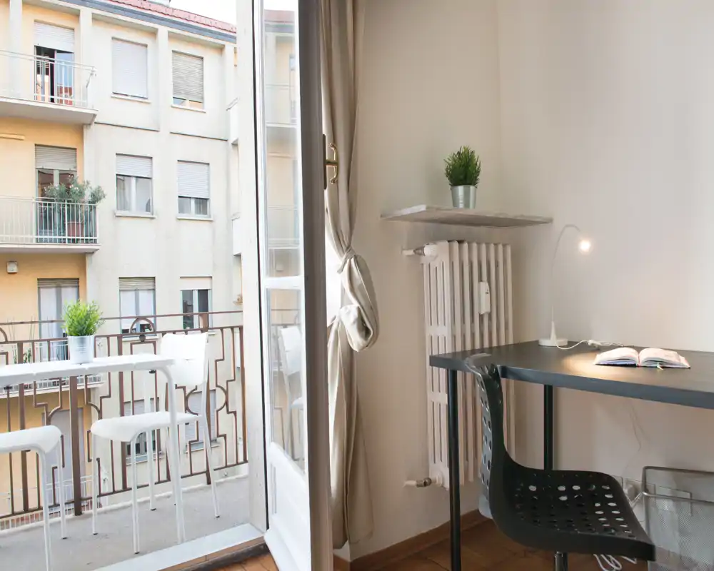Espace de vie moderne et lumineux avec un petit bureau et accès au balcon. La pièce comprend un bureau compact avec lampe, une chaise, des plantes et un coin extérieur visible par la porte ouverte.
