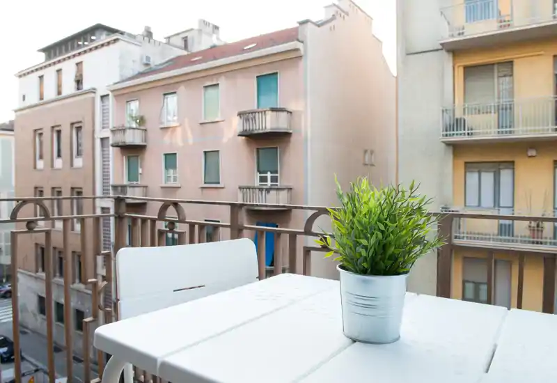 Piccolo balcone soleggiato con tavolo bianco, sedia e una pianta in vaso che si affaccia sugli edifici vicini — ideale per un caffè o una pausa.