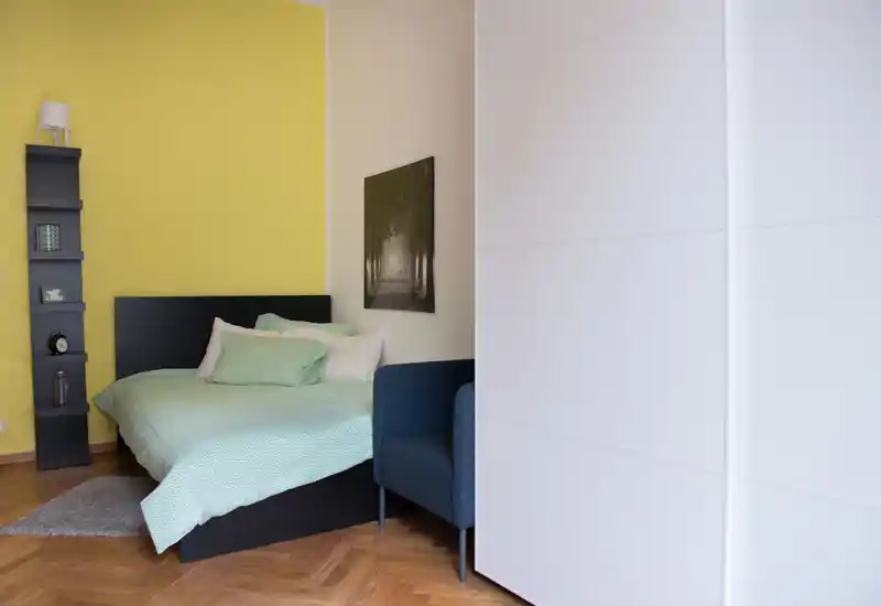 Chambre moderne et lumineuse avec un lit double habillé de linge de lit vert menthe, coussins décoratifs, petit tapis et fauteuil bleu marine; mur accent jaune et armoire blanche pour un espace accueillant.