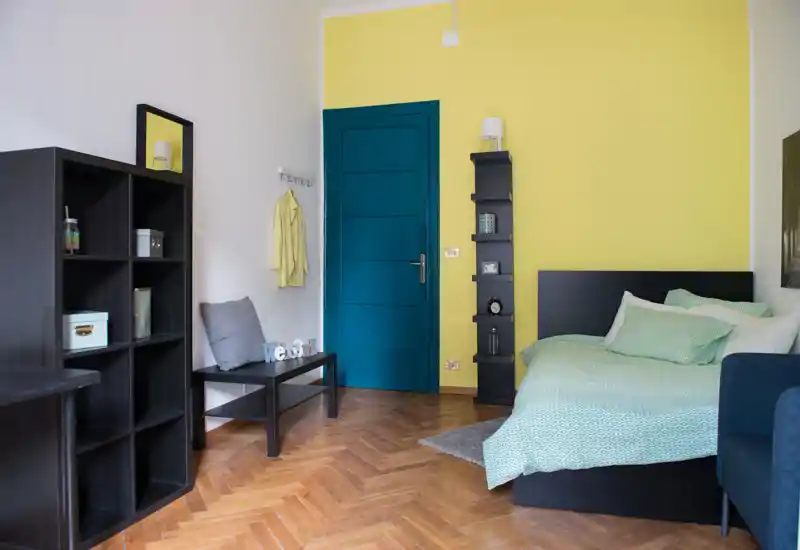 Chambre lumineuse et moderne avec un lit double en linge vert menthe, une tête de lit sombre, un parquet en bois et une porte bleu sarcelle contrastante — étagères et banc complètent l'ambiance cosy.