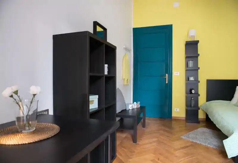 Coin chambre lumineux et moderne avec une porte bleu sarcelle et un mur jaune contrastant. Le lit est visible à droite avec une literie verte, complété par des étagères foncées et un parquet en bois pour une ambiance chaleureuse et contemporaine.