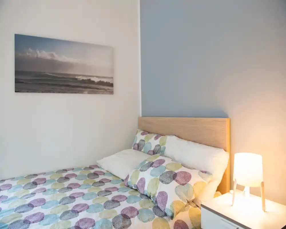 Rincón de dormitorio acogedor y bien iluminado con cabecero de madera, ropa de cama estampada y una lámpara de mesita que aporta calidez.