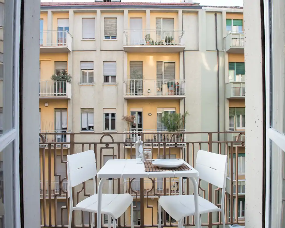 Pequeño balcón soleado con una mesa bistró blanca y dos sillas, con vistas a las fachadas de los edificios vecinos — ideal para café matutino o comidas ligeras.