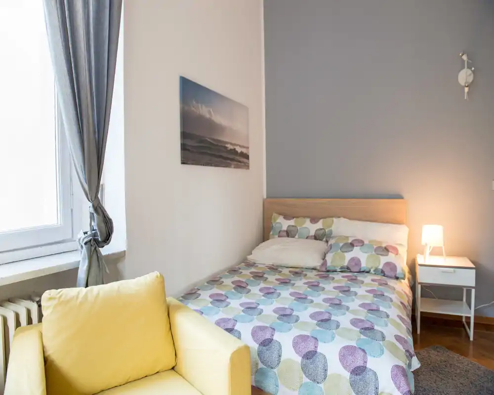 Dormitorio luminoso y ordenado con cama doble estampada, mesita de noche y lámpara cálida. La luz natural de la ventana y la decoración suave generan un ambiente acogedor.