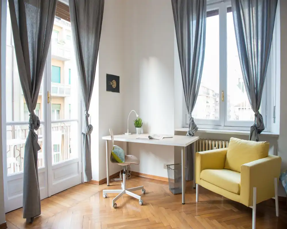 Sala luminosa y con estilo, con grandes ventanas, un escritorio compacto y un cómodo sillón amarillo — ideal para teletrabajar o relajarse con luz natural.