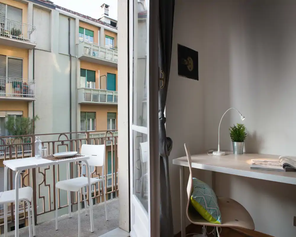 Pequeño balcón amueblado con mesa y taburetes altos con vistas a los edificios vecinos; luz natural y un compacto espacio para comer al aire libre.