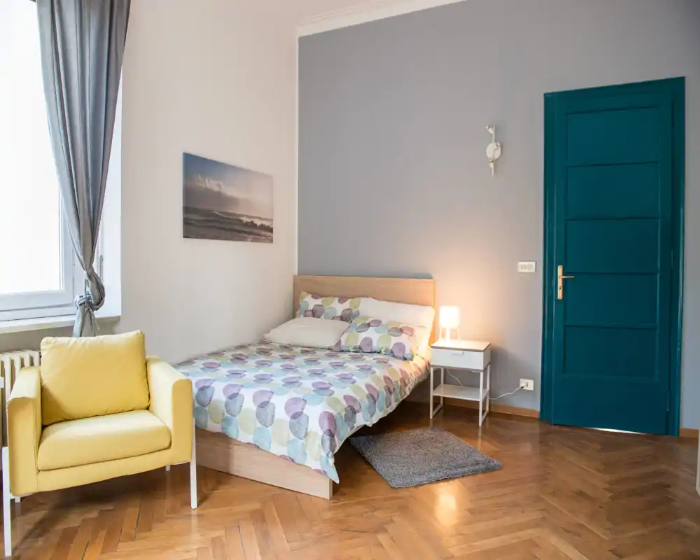 Dormitorio luminoso y con estilo con cama doble, mesita de noche y un cómodo sillón amarillo junto a la ventana. Ropa de cama estampada, lámpara y suelos de parquet aportan una sensación acogedora.