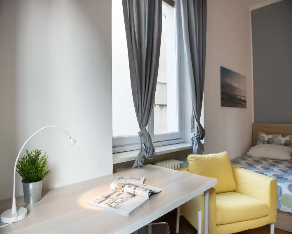 Rincón de dormitorio luminoso y elegante con un cómodo sillón amarillo, un escritorio ordenado con lámpara y revista, y una cama con ropa de cama estampada junto a una gran ventana que deja entrar luz natural suave.