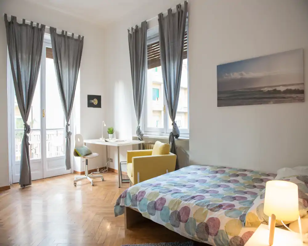 Dormitorio luminoso y acogedor con cama doble, edredón estampado, un sillón cómodo y un pequeño escritorio junto a grandes ventanas que aportan mucha luz natural.