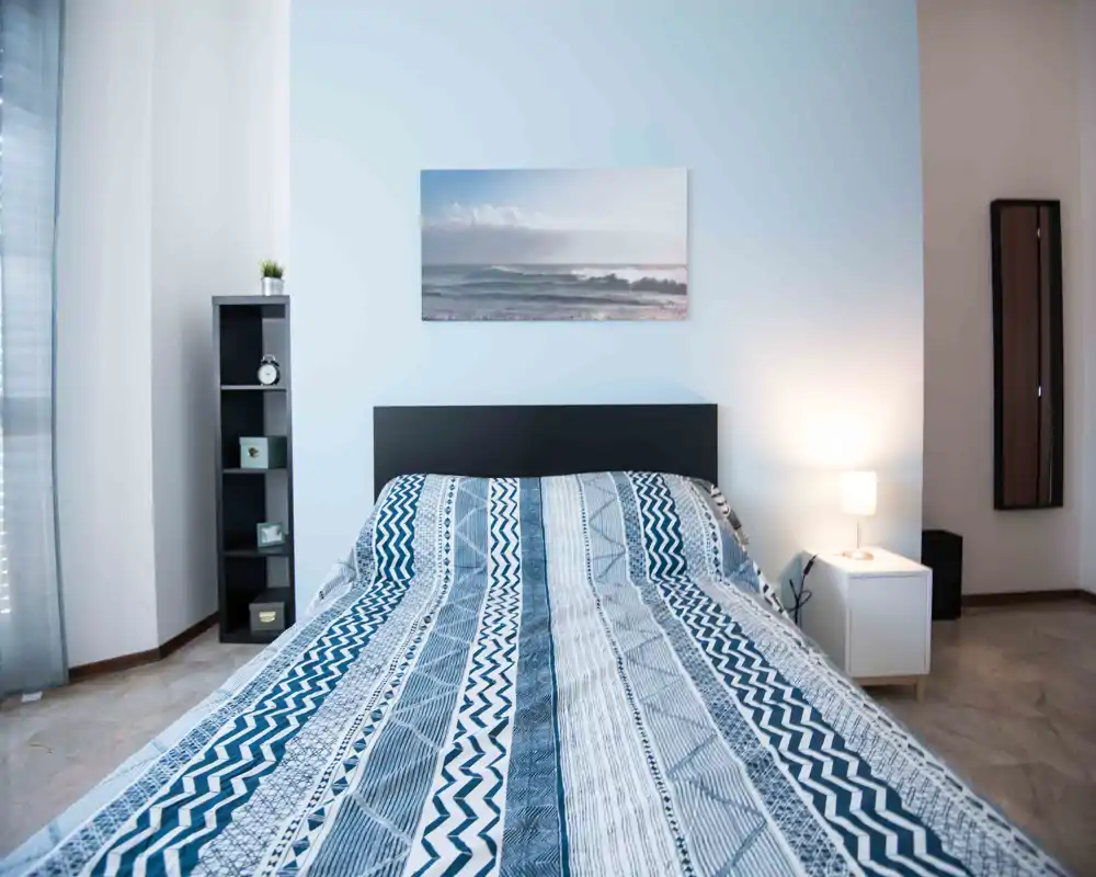 Dormitorio moderno y luminoso con cama doble estampada, cabecero sencillo, lámpara de mesita y decoración ordenada — ideal para anuncios que destaquen confort y estilo minimalista.
