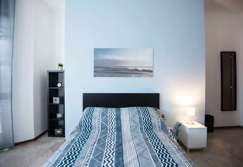 Dormitorio luminoso y ordenado con cama doble estampada, lámpara de noche y estanterías sencillas. Decoración neutra y cuadro costero que aportan un ambiente moderno y relajante.