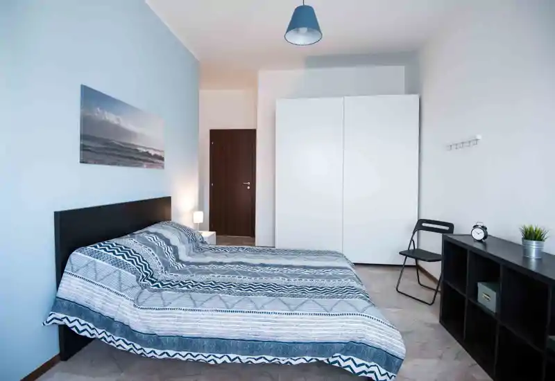 Dormitorio luminoso y ordenado con cama doble, cabecero moderno y ropa de cama estampada, armario blanco y estantería sencilla—ideal para anuncios de alquiler.