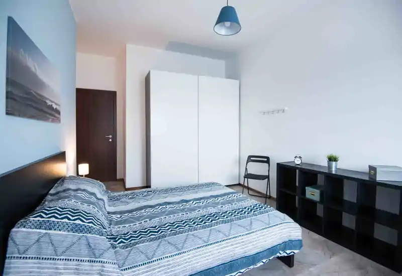 Dormitorio moderno y luminoso con cama doble y ropa de cama azul con estampado, armario blanco, estantería y decoración minimalista — limpio y acogedor para alquileres de corta estancia.