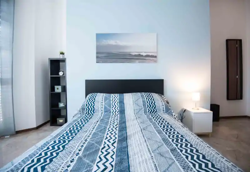 Dormitorio moderno y luminoso con cama doble estampada, cabecero sencillo, lámpara de mesita y decoración ordenada — ideal para anuncios que destaquen confort y estilo minimalista.