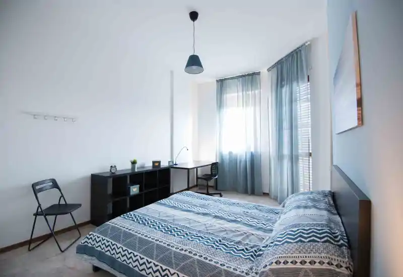 Dormitorio luminoso y minimalista con cama doble con ropa de cama azul estampada, escritorio junto a la ventana y luz natural suave filtrándose por las cortinas.
